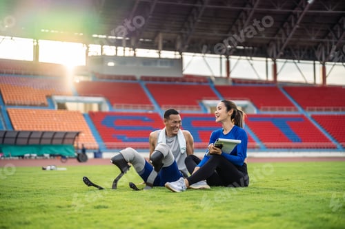 Preview: Asian para-athlete with prosthetic blades and trainer sit in stadium.