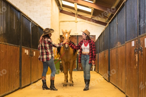 Preview: Two cowgirl women having fun and laughing with a horse in a stable