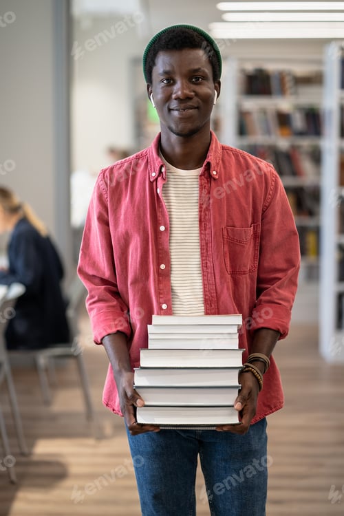 Preview: Portrait of happy African American student guy with pile of books in library