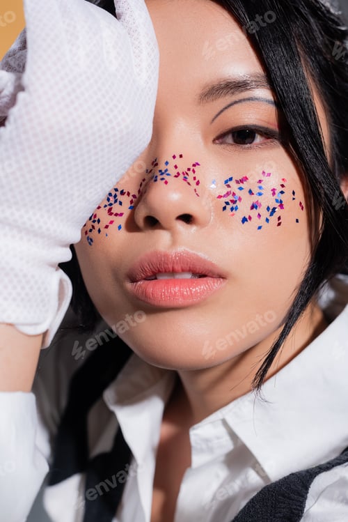 Preview: Portrait of stylish asian model with glitter on face looking at camera isolated on orange