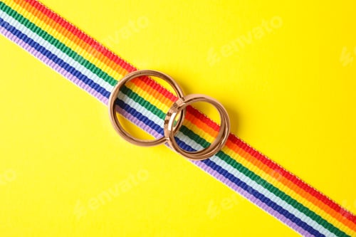 Preview: Wedding rings and rainbow ribbon on color background, top view. Gay symbol