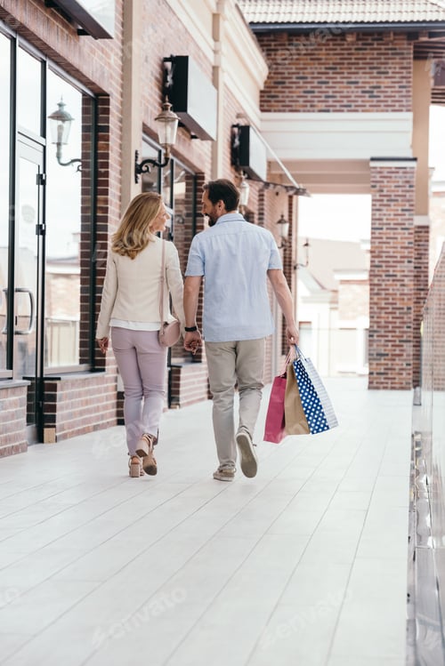 Preview: back view of couple holding hands and walking with shopping bags