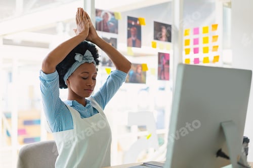 Preview: Woman stretching arms while meditating at desk with computer, coffee mug, sticky notes in workspace