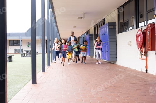 Preview: Diverse male teacher with happy children running in elementary school corridor, copy space