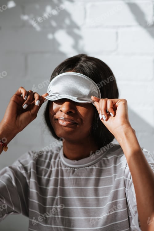 Preview: Happy smiling African female in a white sleeping eye mask sitting on the bed in the morning