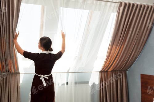 Preview: Housekeeper opening curtains in a bright hotel room during the morning