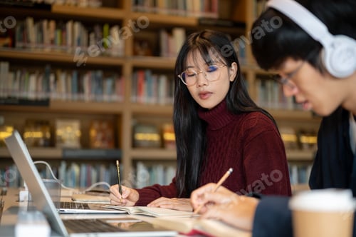 Preview: Glasses asian woman student looking at man friend writing in notebook while sits at library's table.