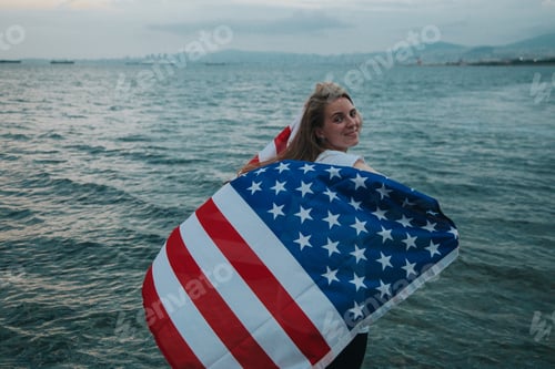 Preview: Young Caucasian girl holding an American Flag in her hands with the sea in the background at sunset