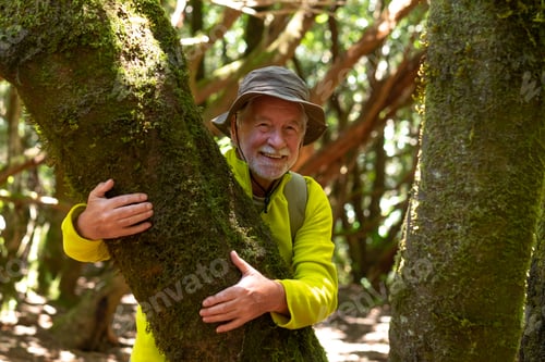 Preview: Senior caucasian man with backpack and cap hiking in the forest hugging a moss covered tree trunk