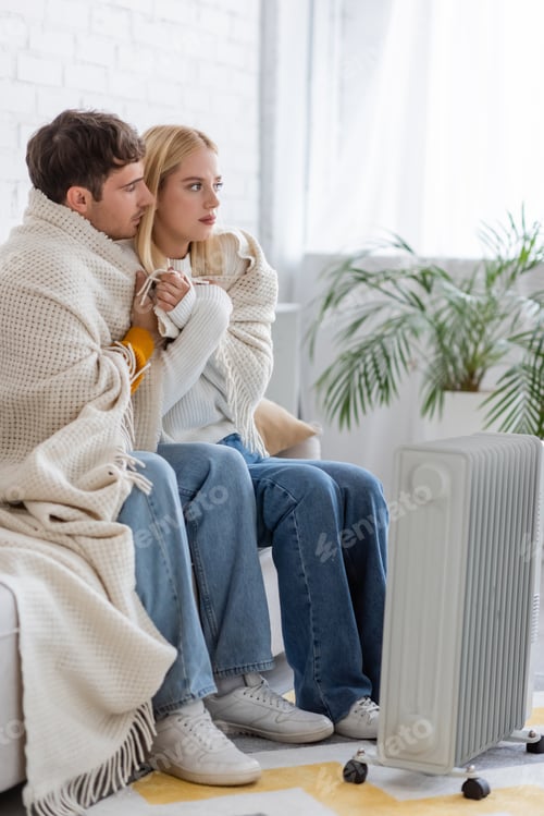 Preview: young couple covered in warm blanket sitting on couch near radiator heater at home