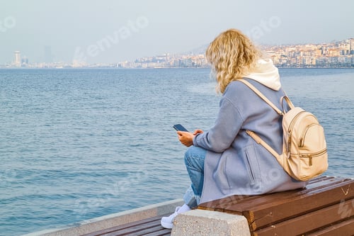 Preview: Blonde woman using her cellphone sitting on the bench on the sea embankment