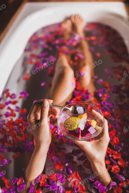 Preview: Woman legs in flower bath in spa. Hands holding plate with fresh fruit salad, view from above