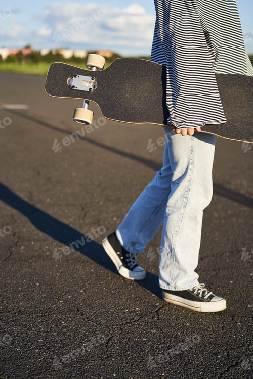 Preview: Cropped shot of teen skater girl, hands holding longboard, walking with skateboard on concrete road
