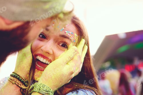 Preview: Portrait of young woman and boyfriend cupping her face with yellow chalked hand at festival