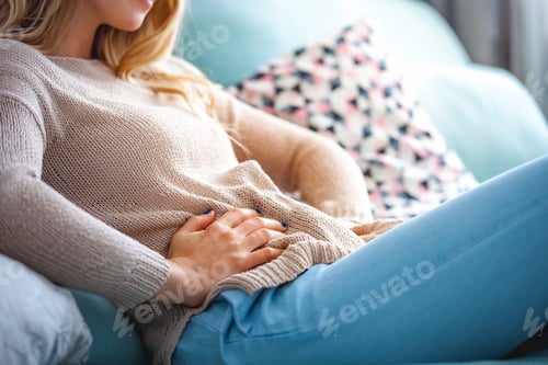 Preview: Young woman suffering from abdominal pain while sitting on sofa at home