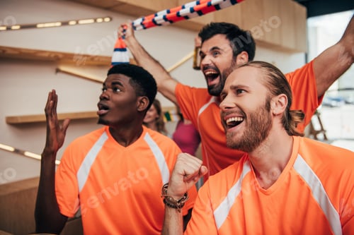 Preview: excited multicultural male football fans in orange t-shirts celebrating and gesturing during watch