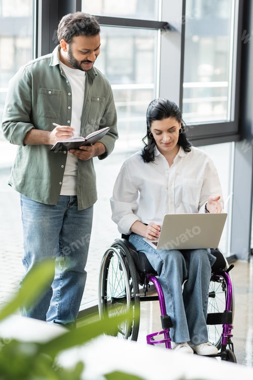Preview: happy indian man taking notes near disabled businesswoman on wheelchair using laptop in office