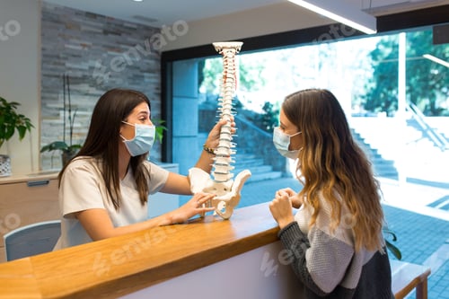 Preview: Physiotherapist explaining the spine to patient wearing surgical face mask