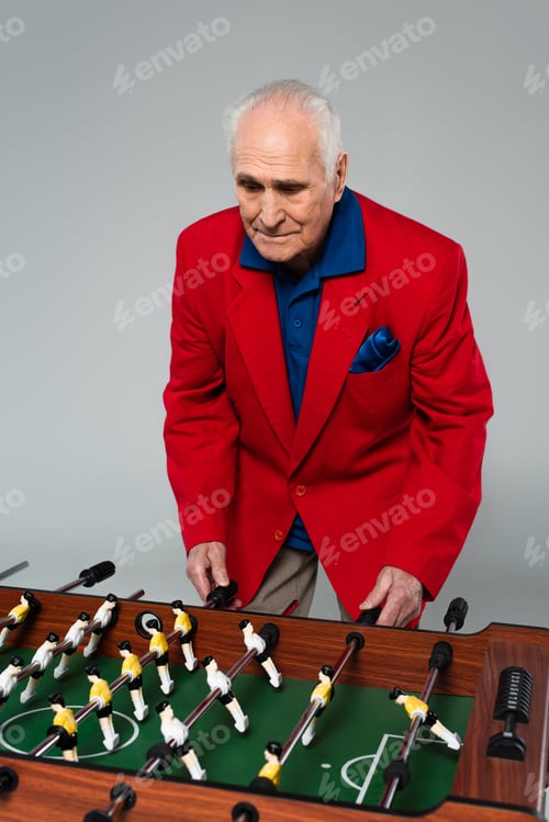 Preview: senior man in red blazer playing table football on grey