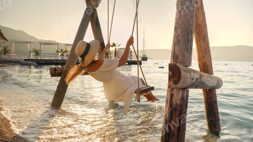 Preview: Young brunette woman in long dress swinging on rope swing at ocean beach bar. Tourism, summertime