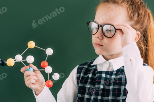 Preview: beautiful little schoolgirl in eyeglasses holding molecular model while standing near chalkboard