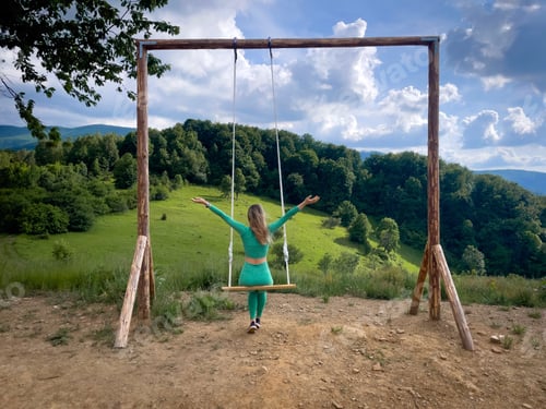 Preview: Rear view of blonde woman sitting on a swing and facing the green mountains on a sunny summer day