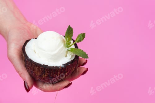 Preview: Vanilla ice cream balls in a fresh coconut half, decorated with mint leaves in your hand on a pink