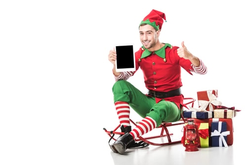 Preview: man in christmas elf costume sitting on sleigh and holding tablet with blank screen isolated on