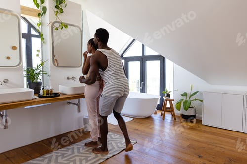 Preview: Happy african american couple brushing teeth and hugging in bathroom