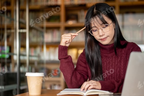 Preview: Glasses asian woman holding pencil and thinking beside laptop and coffee on wooden table in library.