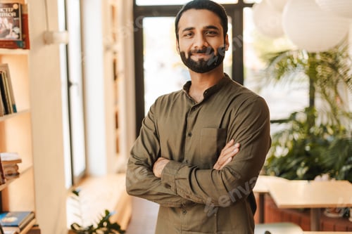 Preview: Confident Indian businessman smiling with crossed arms in office, looking at camera