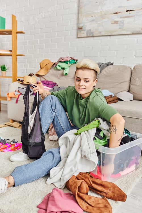Preview: tattooed woman sitting on floor near plastic container and sorting second-hand clothes near couch
