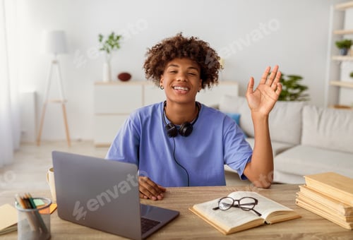 Preview: Portrait of cool Afro teen guy with headphones, laptop and study materials waving at camera, sitting