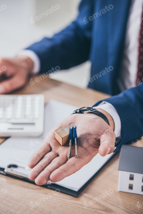 Preview: partial view of businessman at workplace showing keys near house model, clipboard and calculator