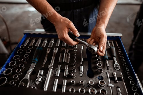 Preview: Man holds ratchet wrench over a toolbox filled with various tools in a workshop setting