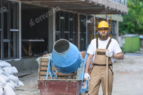 Preview: young builder in hardhat and protective googles standing with shovel near concrete mixer at