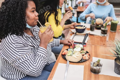 Preview: Young multiracial friends having breakfast in restaurant outdoors with masks under chins