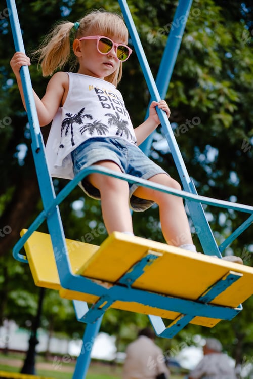Preview: Stylish little girl in pink sunglasses swinging on playground