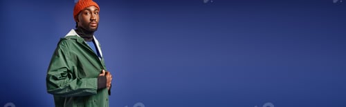 Preview: bearded african american man in knitted hat and winter attire looking at camera on blue, banner