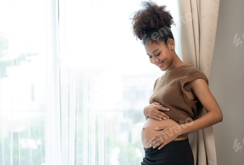 Preview: Happy African American pregnant woman touching belly and standing beside windows in bedroom at home