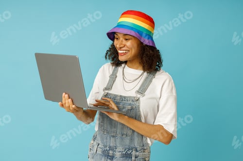 Visualização: Jovem freelancer positiva em boné de arco-íris está usando laptop em fundo azul de estúdio