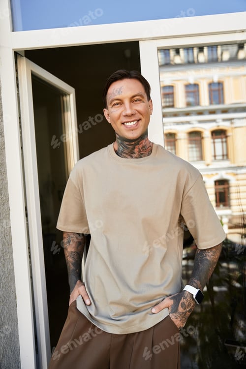 Preview: A young man smiles confidently at home, showcasing his tattoos and relaxed style in daylight