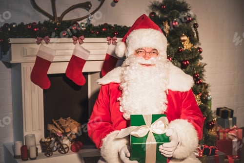 Preview: santa claus holding christmas gift and looking at camera while sitting near fireplace