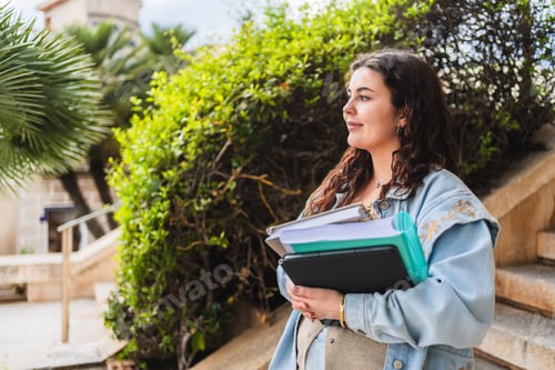 Preview: Smiling university student holding books and looking away on campus
