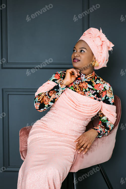 Preview: Portrait of beautiful African-American female model in pink ethnic dress and headwrap, sitting on
