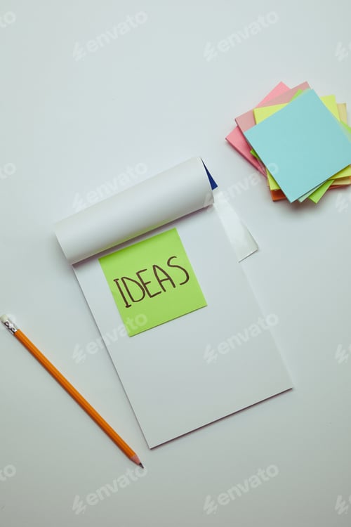 Preview: top view of paper sticker with word ideas in notebook, pencil and pile of note papers on white