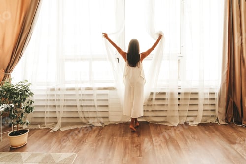 Preview: Young woman stands by large window in silk white gown after waking up at home