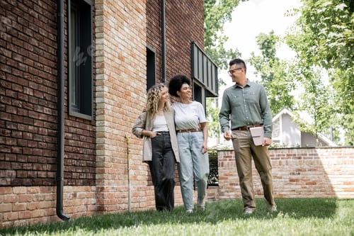 Aperçu: full length of real estate agent and multiethnic lesbian couple walking and talking near new cottage