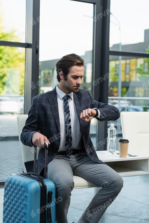 Preview: worried man looking at wristwatch while sitting in departure lounge