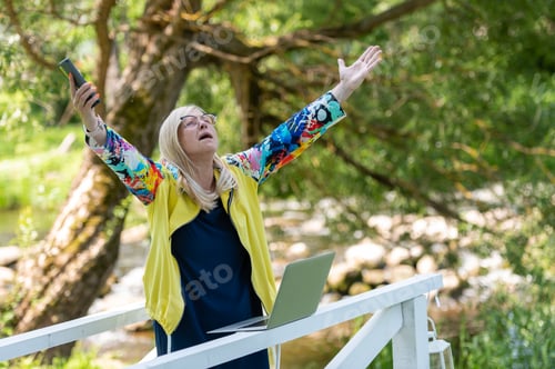 Preview: Desperate angry business woman shouting in anger with hands raised in the air, using phone and lapto